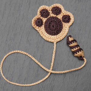 Può includere: Un segnalibro a forma di impronta di zampa all'uncinetto con una coda marrone. L'impronta della zampa è beige con dettagli marroni e la coda è marrone e beige.