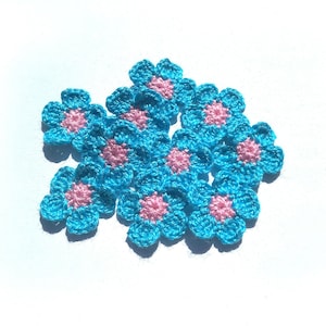 May include: A cluster of small, crocheted flowers. Each flower features light blue petals and a pink centre. The flowers are arranged on a white surface, giving a delicate, handmade appearance.