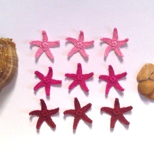 May include: A seashell with a brown and tan pattern, surrounded by pink and red crocheted starfish. There are also several small brown stones.