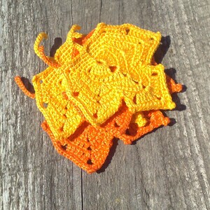 May include: A collection of handmade crochet squares in shades of yellow and orange. The squares have a scalloped edge and are arranged on a weathered wooden surface. The crochet work appears to be tightly woven, with a textured appearance.