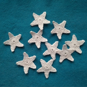 May include: Nine white crocheted star appliques are scattered on a teal blue background. Each star has five points and a small hole in the center. The stars are handmade and appear to be made of cotton yarn.