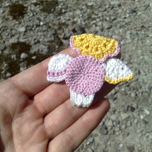 Puede incluir: Una flor de ganchillo hecha a mano en tonos amarillo, rosa y blanco. La flor se sostiene en una mano, mostrando el intrincado trabajo de puntadas y el dise&ntilde;o delicado. Los p&eacute;talos y el centro de la flor est&aacute;n claramente definidos, con un toque de verde en la base.