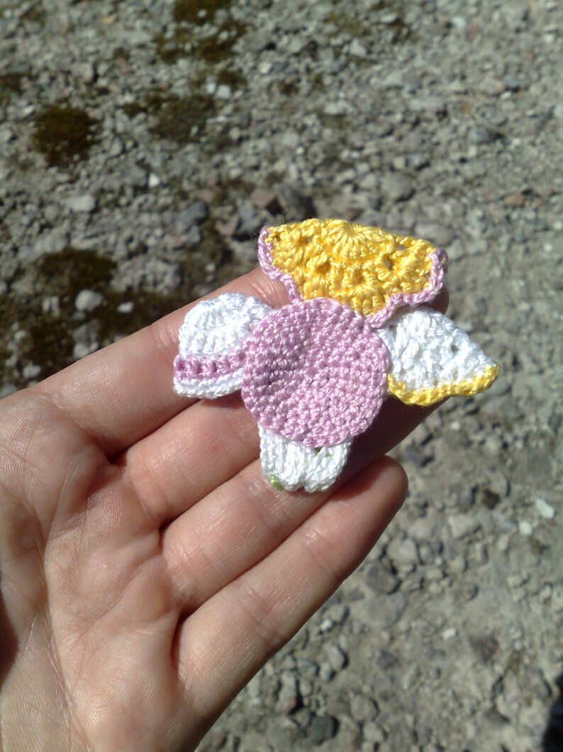 Puede incluir: Una flor de ganchillo hecha a mano en tonos amarillo, rosa y blanco. La flor se sostiene en una mano, mostrando el intrincado trabajo de puntadas y el dise&ntilde;o delicado. Los p&eacute;talos y el centro de la flor est&aacute;n claramente definidos, con un toque de verde en la base.