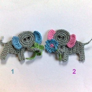 May include: Two crocheted elephant appliques. One has blue ears and a green flower, the other has pink ears and a blue flower. Both are grey with black eyes and a curled trunk. The appliques are handmade.