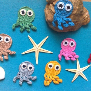 Può includere: Una collezione di ornamenti a forma di polpo all'uncinetto fatti a mano in vari colori, tra cui verde, marrone, blu, rosa, grigio, giallo e rosso. I polpi hanno grandi occhi a palla. La scena comprende stelle marine e conchiglie su sfondo blu.
