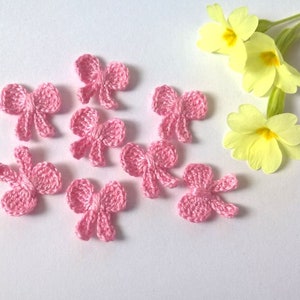 Pode incluir: Uma coleção de pequenos laços rosa de crochê feitos à mão sobre uma superfície branca. Também estão presentes flores amarelas de prímulas. Os laços são provavelmente para artesanato ou decoração.