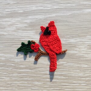May include: A handmade crochet cardinal bird in vibrant red, perched on a brown branch with green leaves and red berries. The bird has a black head and is set against a light wood grain background. This decorative item is perfect for nature lovers.