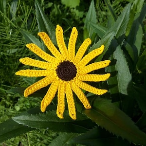 May include: A handmade crochet flower with yellow petals and a dark brown center. The flower is set against a backdrop of green leaves, creating a natural and vibrant contrast. This craft item is ideal for decorative purposes.