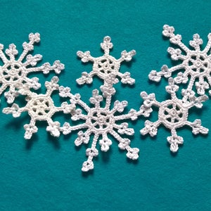May include: Six white, crocheted snowflakes arranged on a teal background. Each snowflake has a detailed, symmetrical design with multiple points. The snowflakes are made of a soft, textured yarn, perfect for winter decorations.
