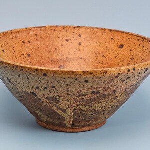 May include: A ceramic bowl with a wide, flared rim and a tapered base. The exterior and interior feature a speckled, mottled texture with earthy tones of tan, brown, and dark orange, showcasing a variegated glaze finish across the entire surface.