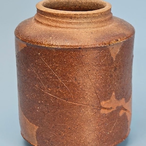 May include: A cylindrical ceramic jar with a textured, reddish-brown finish. The exterior surface features a subtle, embossed leaf pattern that wraps around the body. The vessel has a short, thick neck and a wide, open circular mouth.