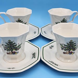 Puede incluir: Cuatro tazas y platillos blancos con un diseño de árbol de Navidad. Las tazas tienen una forma angular única y los platillos son octogonales con un borde verde. Sobre un fondo azul.