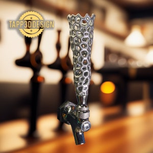 May include: A silver beer tap handle with a honeycomb pattern. The tap handle has a textured, open design. The tap is connected to a chrome faucet with a lever. The background is blurred, suggesting a bar. The text "TAPP3D DESIGN" is in the upper left.