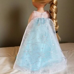 May include: A doll wearing a light blue dress with a white tulle overlay and silver snowflake and swirl designs. The doll has long blonde hair in a braid.