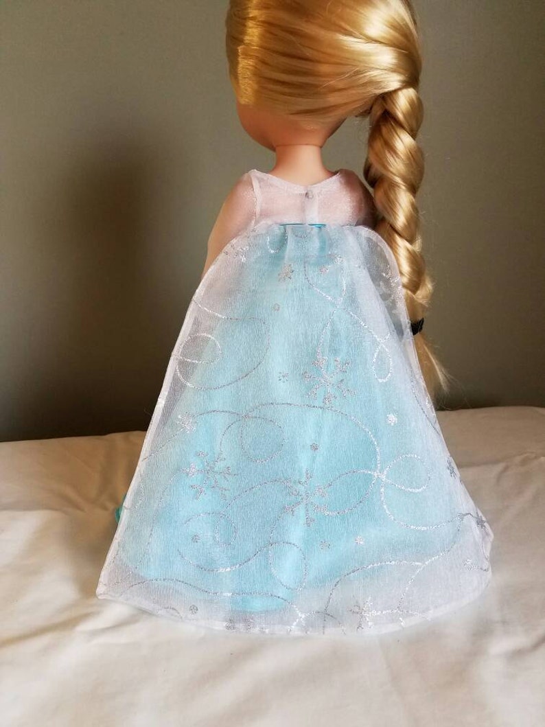 May include: A doll wearing a light blue dress with a white tulle overlay and silver snowflake and swirl designs. The doll has long blonde hair in a braid.