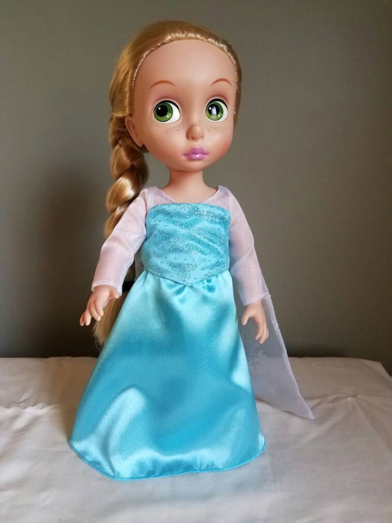 May include: A blonde doll wearing a light blue dress with a sparkly bodice and a sheer white cape. The doll has green eyes and pink lips.