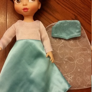 May include: A doll wearing a light blue dress with a white bodice and long sleeves. The doll has brown hair and a light complexion. The doll is wearing a light blue cape with a sparkly design.