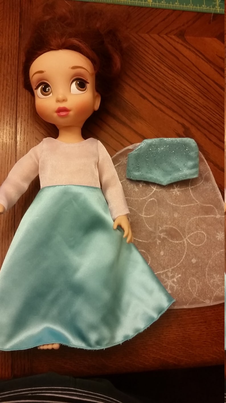 May include: A doll wearing a light blue dress with a white bodice and long sleeves. The doll has brown hair and a light complexion. The doll is wearing a light blue cape with a sparkly design.