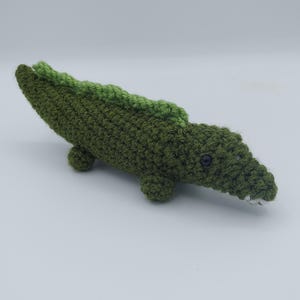 May include: A handmade crochet alligator toy in shades of green. The body is a darker green, with a lighter green ridge along the back. The alligator has small, round feet, black eyes, and white teeth.