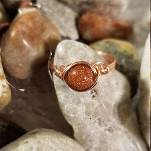 May include: A copper wire wrapped ring with a round, orange sunstone gemstone.