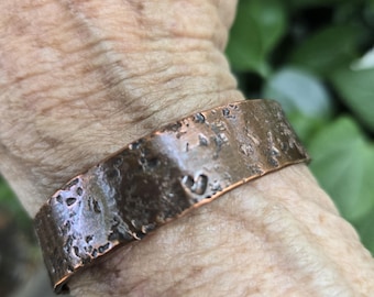 May include: A wide copper cuff bracelet with a dark, oxidized finish and a heavily textured, hammered surface. The metal band features an uneven, rugged appearance with deep indentations and a metallic brown patina across the entire exterior.