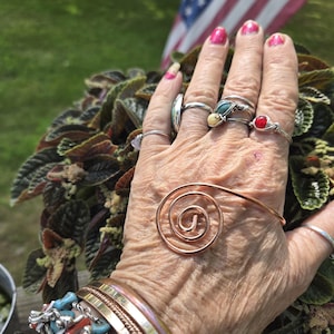 May include: A hand adorned with various rings and bracelets. Rings include silver bands with gemstones and wire-wrapped designs. A copper spiral bracelet is also visible. The bracelets feature turquoise and silver accents. The background includes a plant and an American flag.