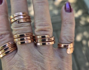 May include: A collection of copper-colored metal rings worn on four fingers. Each ring features a multi-layered wrap design with a smooth, polished finish. The bands are constructed from flat, thin strips of metal that coil around the finger.