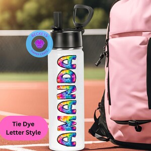 May include: White water bottle with a tie-dye design spelling out the name "Amanda". The bottle has a black lid with a carrying handle and a straw.