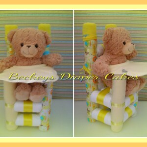 Op de afbeelding: Een gele en witte luiercake in de vorm van een hoge stoel met een bruine teddybeer die erin zit. De cake is versierd met geel en wit lint en een wit slabbetje. De cake is gemaakt van luiers, babydoekjes en andere babybenodigdheden.