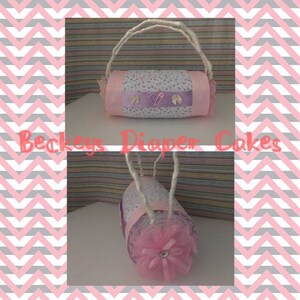 May include: A pink and purple diaper cake with a white handle and a pink bow. The cake is decorated with white polka dots and a purple ribbon. The text "Beckeys Diaper Cakes" is visible in the image.
