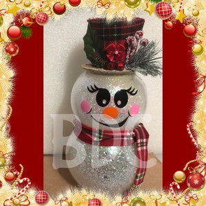 Peut inclure: Une figurine de bonhomme de neige en paillettes blanches avec un chapeau en tartan rouge et noir, un n&oelig;ud rouge et une &eacute;charpe en tartan rouge et noir. Le bonhomme de neige a un visage noir et blanc avec un sourire noir, des yeux noirs et un nez orange.