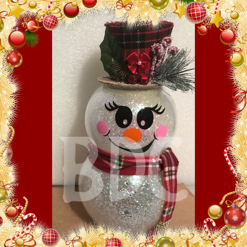Peut inclure: Une figurine de bonhomme de neige en paillettes blanches avec un chapeau en tartan rouge et noir, un n&oelig;ud rouge et une &eacute;charpe en tartan rouge et noir. Le bonhomme de neige a un visage noir et blanc avec un sourire noir, des yeux noirs et un nez orange.