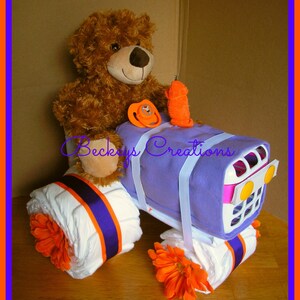 May include: A purple and white diaper cake shaped like a tractor with orange flowers and a brown teddy bear sitting on top. The cake is decorated with white ribbon and a pacifier.