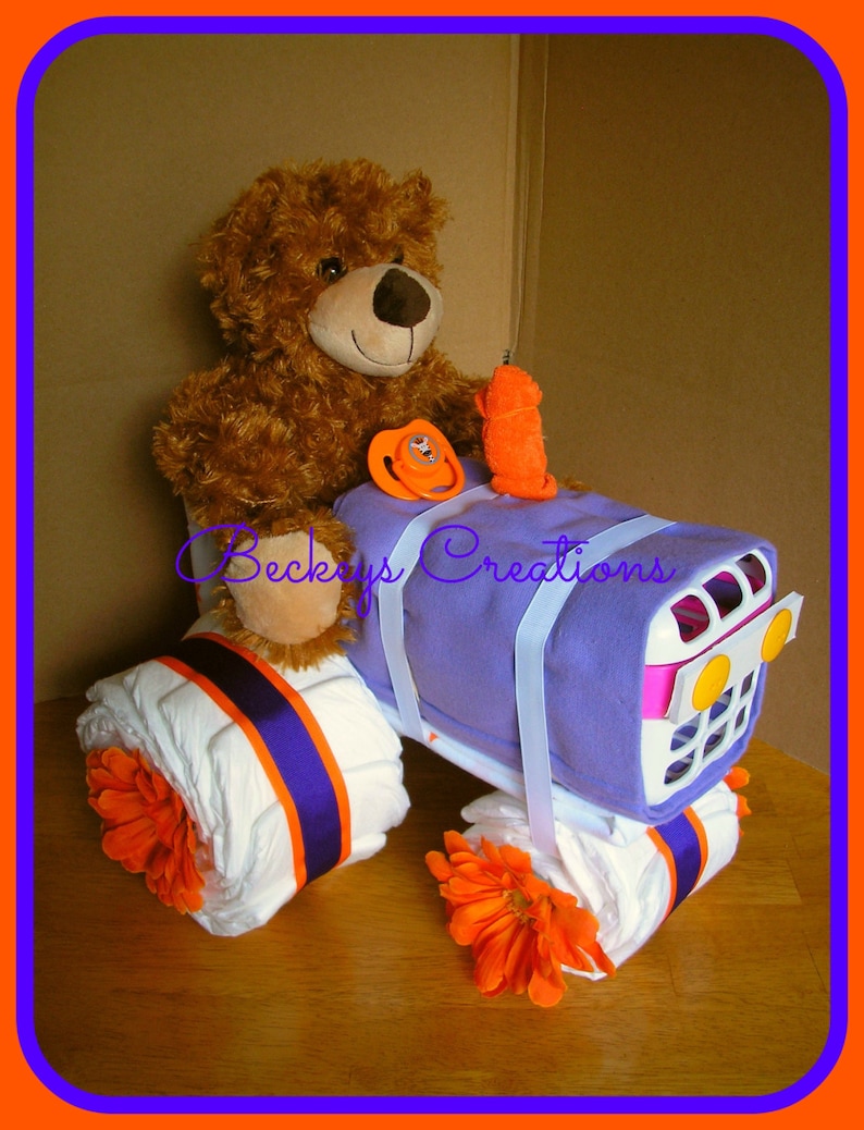 May include: A purple and white diaper cake shaped like a tractor with orange flowers and a brown teddy bear sitting on top. The cake is decorated with white ribbon and a pacifier.