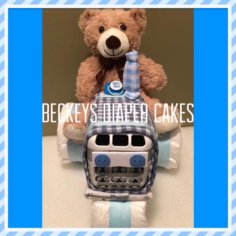 May include: A blue and white checkered diaper cake shaped like a bus with a brown teddy bear sitting on top. The teddy bear is wearing a blue and white checkered bow tie. The cake is decorated with a blue and white pacifier and a blue and white "It's a Boy" sign.