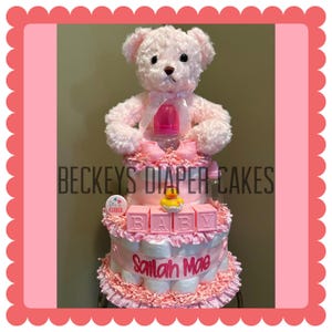May include: A pink diaper cake with a teddy bear topper, baby bottle, and baby shoes. The cake has three tiers with the text "BECKEYS DIAPER CAKES", "BABY", and "Sailah Mae". A rubber ducky sits on the middle tier.