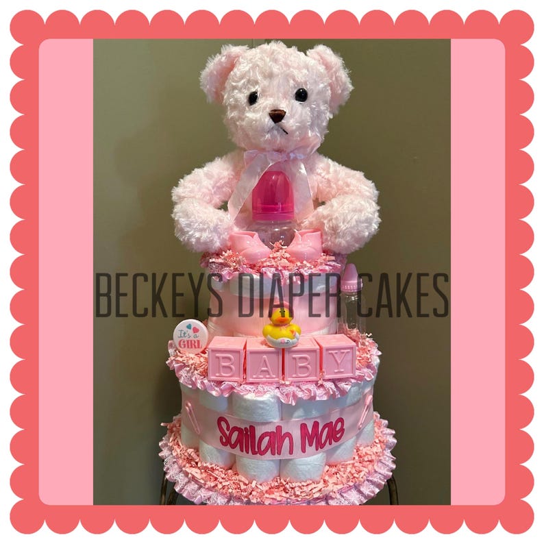 May include: A pink diaper cake with a teddy bear topper, baby bottle, and baby shoes. The cake has three tiers with the text "BECKEYS DIAPER CAKES", "BABY", and "Sailah Mae". A rubber ducky sits on the middle tier.