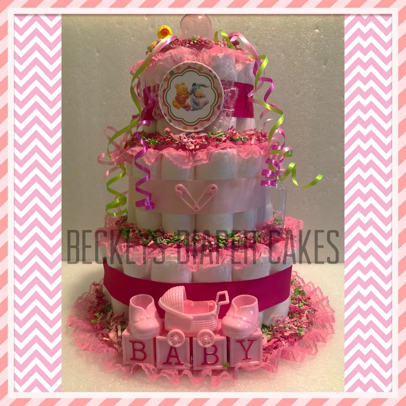 May include: A three-tiered diaper cake decorated with pink and green ribbons, a Winnie the Pooh and Tigger character topper, and a pink baby carriage with pink baby shoes. The bottom tier has a pink ribbon and the words "BABY" spelled out in pink blocks.