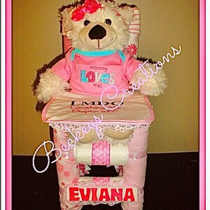 Op de afbeelding: Een roze en witte luiercake met een witte teddybeer die een roze shirt draagt ​​met de tekst "mama houdt van me!" De cake is versierd met roze en witte stippen en kant. De cake is gelabeld met "LMDO" en "Eviana".