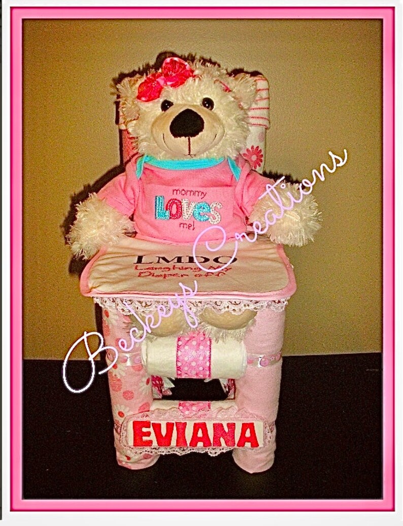 Op de afbeelding: Een roze en witte luiercake met een witte teddybeer die een roze shirt draagt ​​met de tekst "mama houdt van me!" De cake is versierd met roze en witte stippen en kant. De cake is gelabeld met "LMDO" en "Eviana".