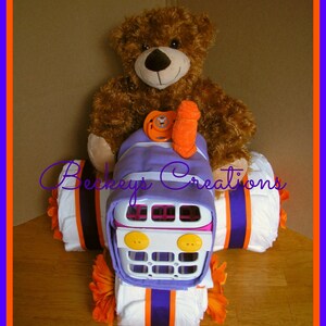 May include: A purple and white diaper cake shaped like a tractor with a brown teddy bear sitting on top. The tractor has a white plastic grill and yellow buttons. The diaper cake is decorated with orange and purple ribbon.