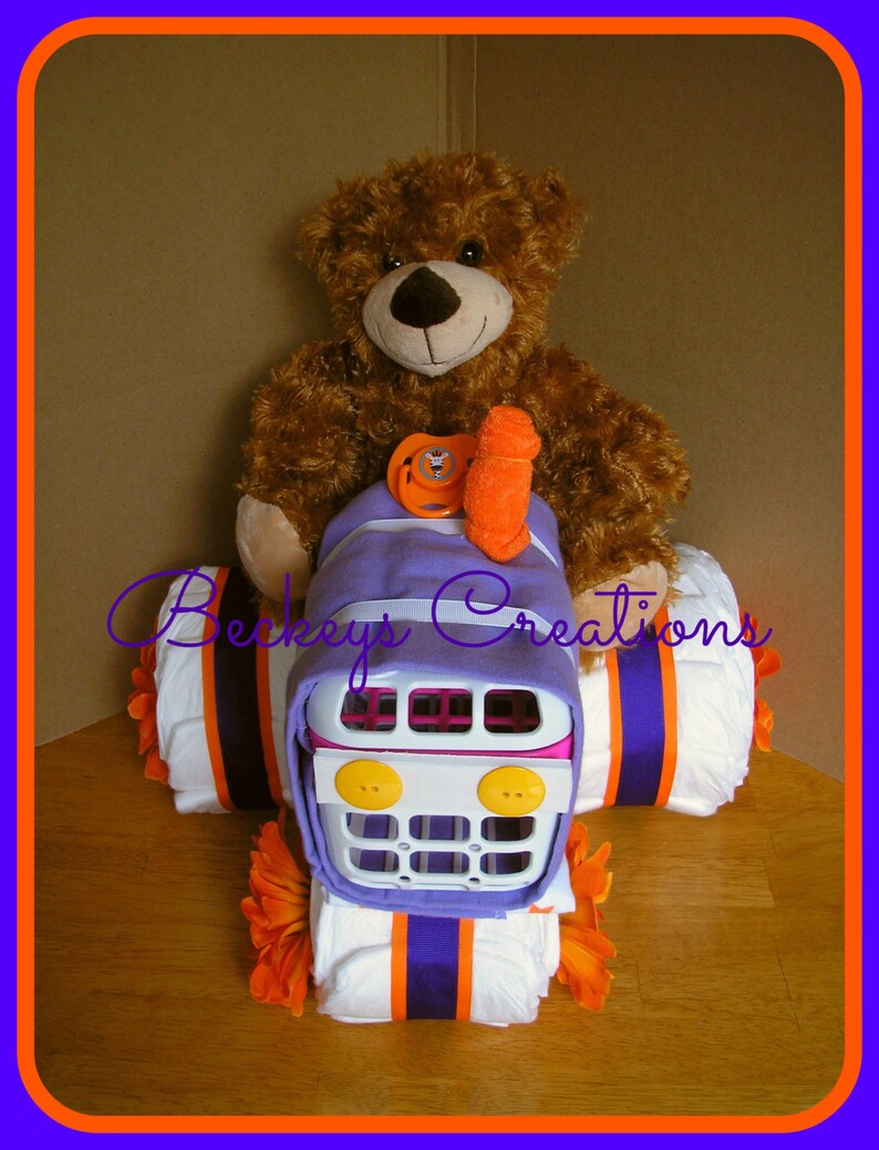 May include: A purple and white diaper cake shaped like a tractor with a brown teddy bear sitting on top. The tractor has a white plastic grill and yellow buttons. The diaper cake is decorated with orange and purple ribbon.