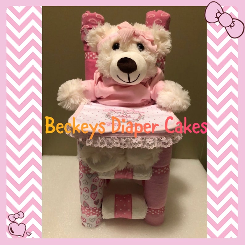 Op de afbeelding: Een roze en witte luiercake met een teddybeer die er bovenop zit. De cake is versierd met hartjes en een roze lint. De teddybeer draagt een roze jurk en heeft een roze strik op zijn hoofd. De cake is gelabeld met "Beckeys Diaper Cakes".