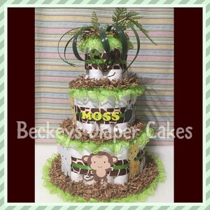 Op de afbeelding: Een drielaagse luiercake versierd met groene en bruine dierenprintstof, groen lint en een groene kantversiering. De cake is bedekt met een groene, bladerrijke plant en een bruin-wit zebrabeeldje. De middelste laag heeft een geel naambordje met de tekst "Moss". De onderste laag is versierd met een bruin aapbeeldje.