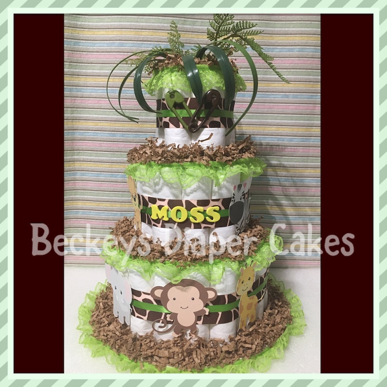Op de afbeelding: Een drielaagse luiercake versierd met groene en bruine dierenprintstof, groen lint en een groene kantversiering. De cake is bedekt met een groene, bladerrijke plant en een bruin-wit zebrabeeldje. De middelste laag heeft een geel naambordje met de tekst "Moss". De onderste laag is versierd met een bruin aapbeeldje.