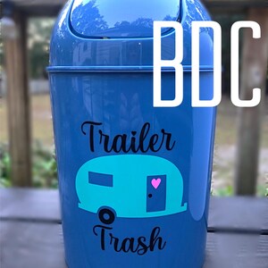 以下が含まれることがあります： 白い蓋と「Trailer Trash」という黒いステッカーが付いた青いプラスチック製のゴミ箱。ターコイズブルーのキャンピングカーのシルエットとドアにピンクのハートが描かれています。
