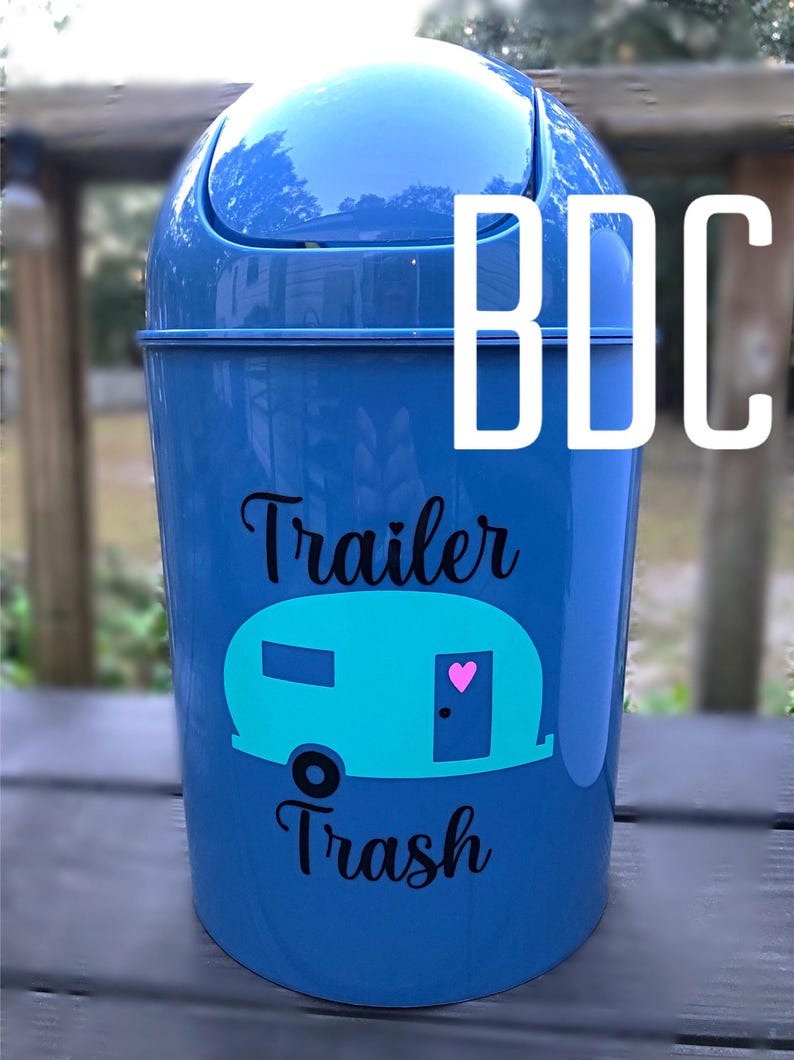 以下が含まれることがあります： 白い蓋と「Trailer Trash」という黒いステッカーが付いた青いプラスチック製のゴミ箱。ターコイズブルーのキャンピングカーのシルエットとドアにピンクのハートが描かれています。