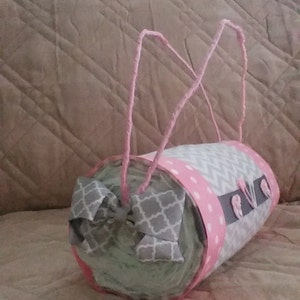 May include: A grey and pink nappy cake with a grey and white bow. The cake is decorated with pink polka dots and grey chevron patterns. The cake is wrapped in pink ribbon and has a grey and white bow on top.