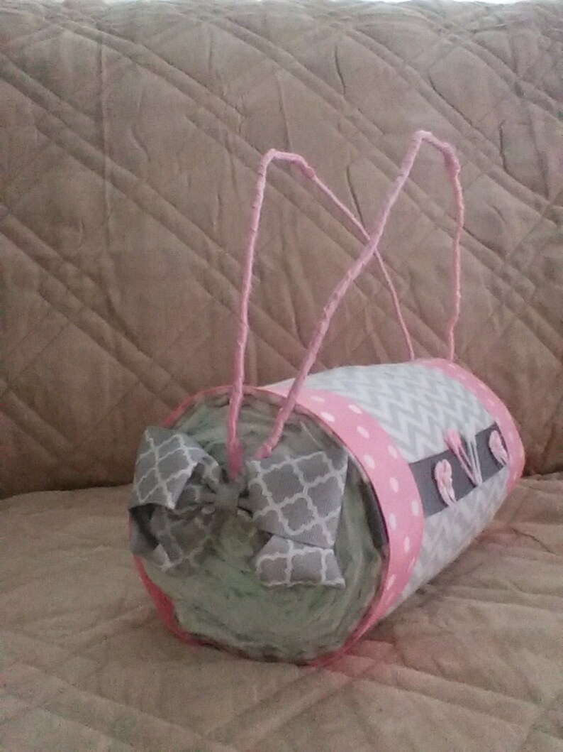 May include: A grey and pink nappy cake with a grey and white bow. The cake is decorated with pink polka dots and grey chevron patterns. The cake is wrapped in pink ribbon and has a grey and white bow on top.