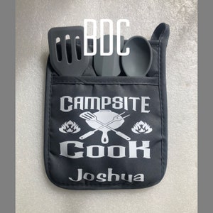 May include: Black oven mitt with the text "Campsite Cook Joshua" in white. The mitt has three pockets for holding utensils, and a spatula, spoon, and turner are shown in the pockets.